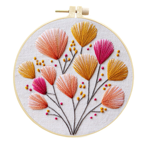 A circular embroidery hoop holding a floral needlepoint or embroidery design on white fabric. The design features stylized flowers in orange, pink, and mustard yellow with thin dark stems and small berry-like accents, photographed against a plain white background.