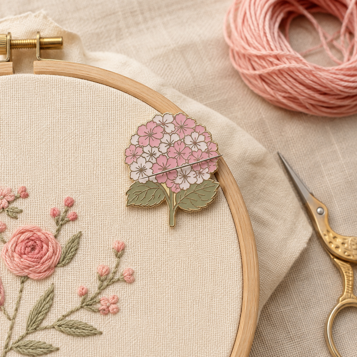A close-up of a wooden embroidery hoop holding cream fabric with pink and white floral stitching. A pink and white flower-shaped pin or charm rests on the hoop, with a pink thread ball, gold scissors, and beige fabric in the background.