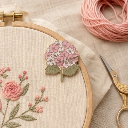 A close-up of a wooden embroidery hoop holding cream fabric with pink and white floral stitching. A pink and white flower-shaped pin or charm rests on the hoop, with a pink thread ball, gold scissors, and beige fabric in the background.
