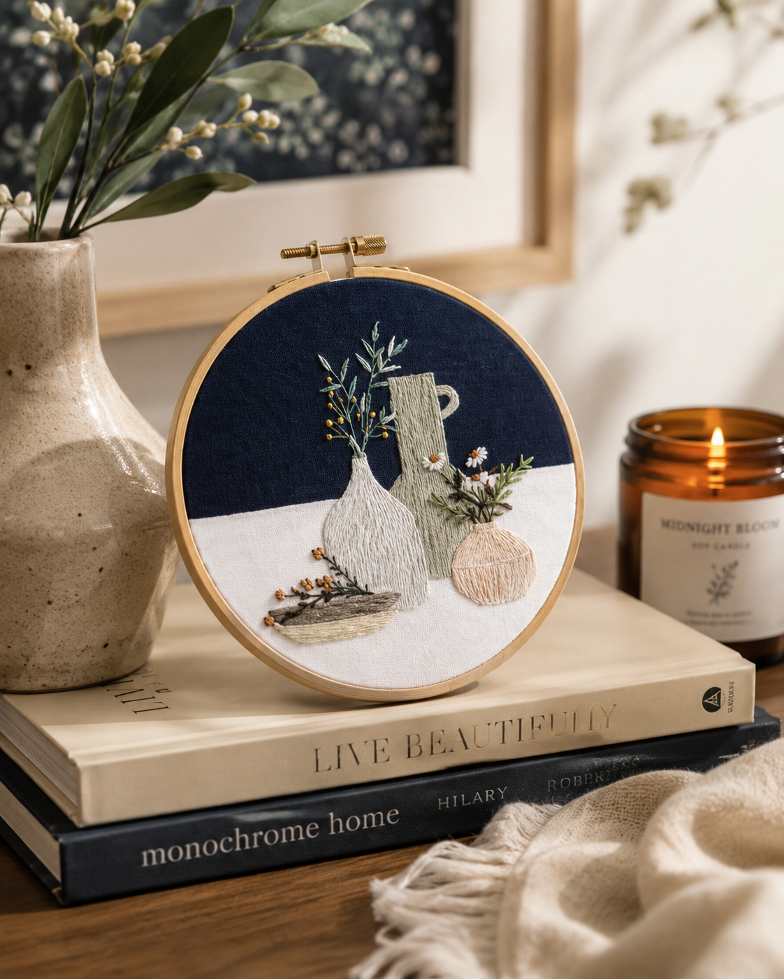 Embroidery hoop with a nature-themed design on a stack of books with a candle and vase in the background.
